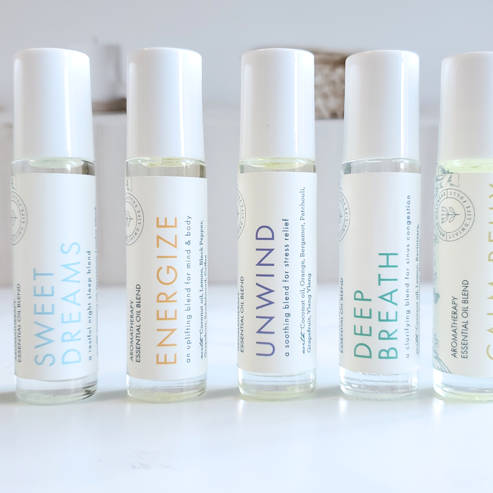 A series of six clear glass roll-on bottles with white labels, each indicating a different essential oil blend name such as 'Energize' and 'Unwind'. The bottles are filled with jojoba oil base and the essential oil blend, and are displayed against a neutral background.