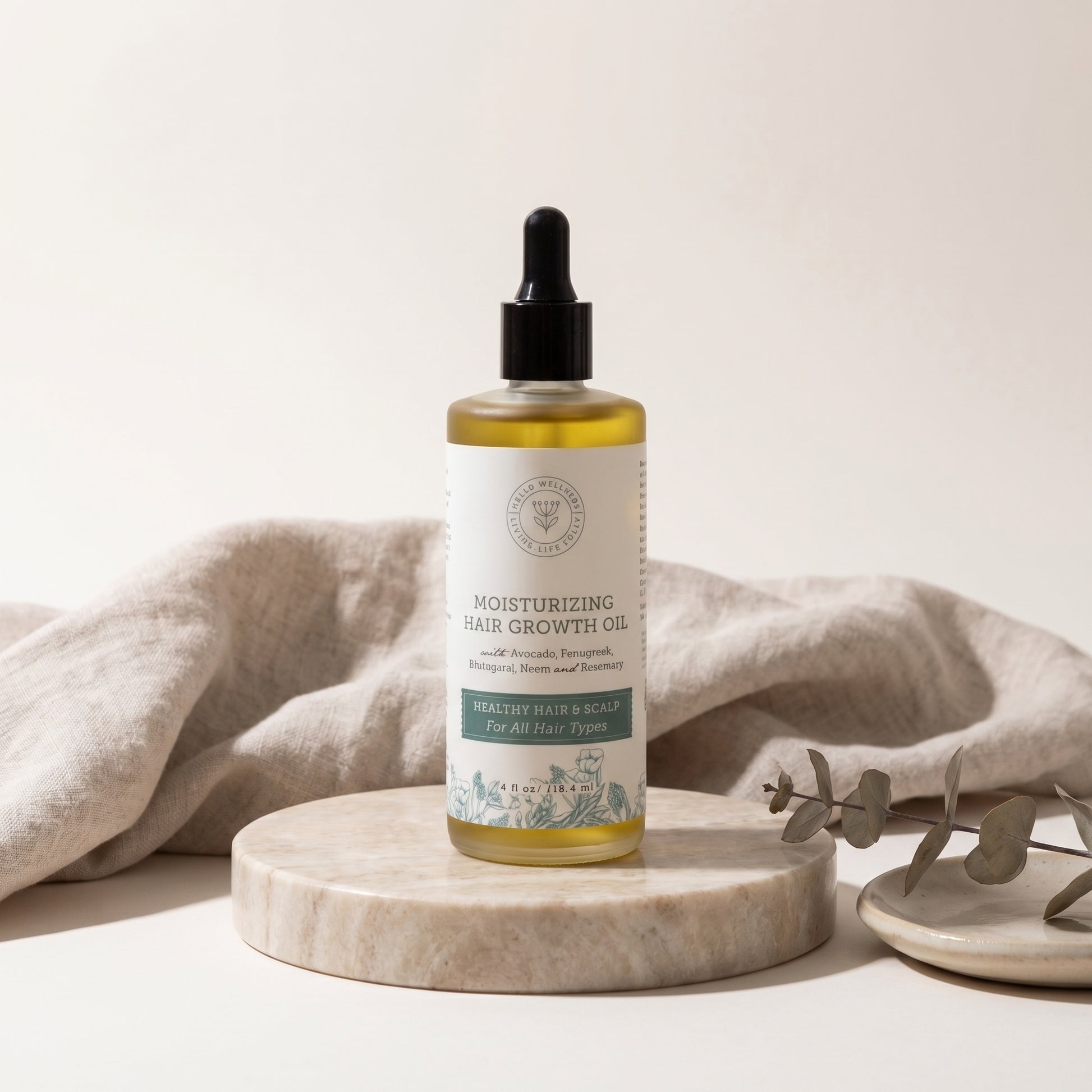 Bottle of moisturizing hair growth oil on a marble surface with a beige cloth and eucalyptus leaves in the background.