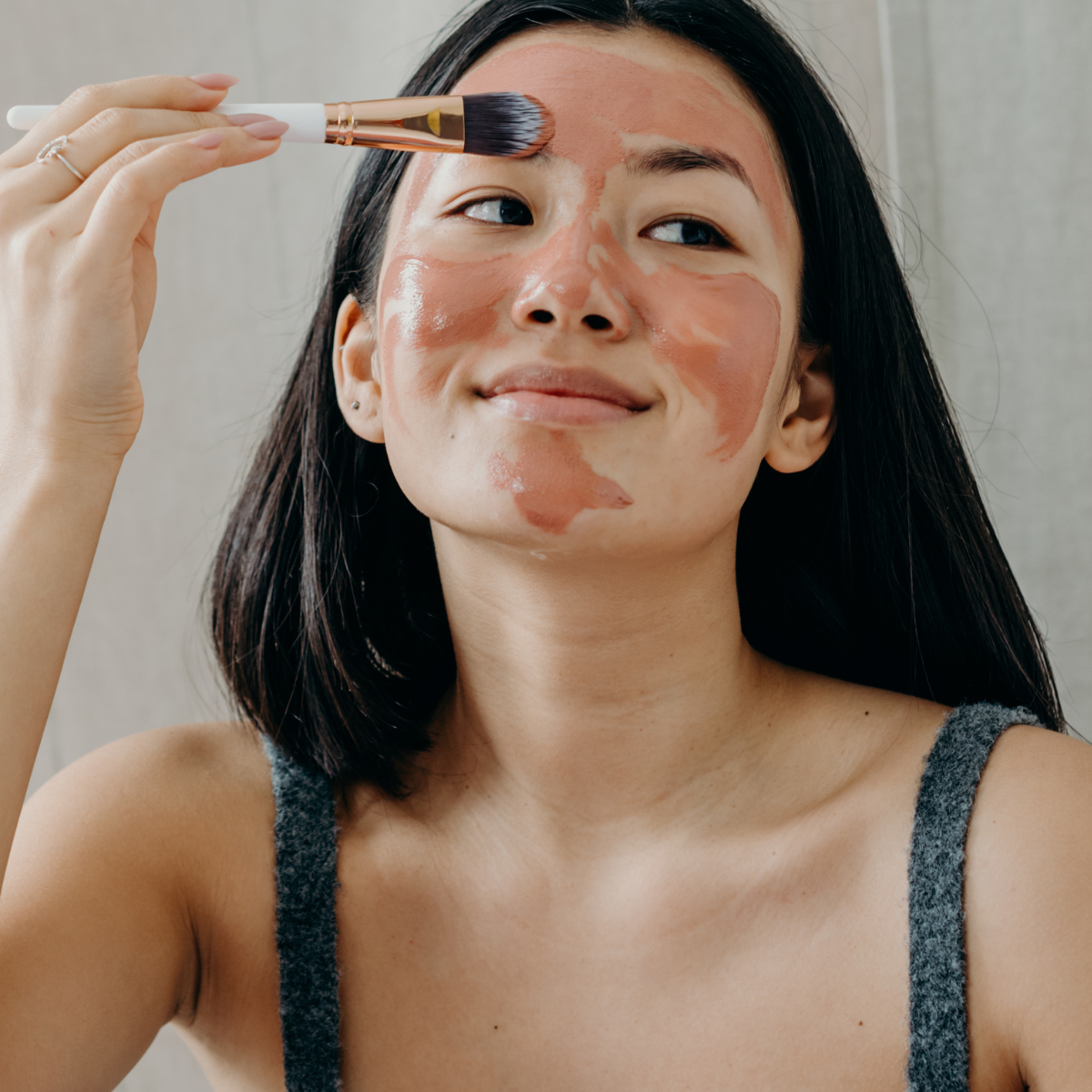 Woman applying a pink clay facial mask to her forehead with a brush as part of her skincare routine.
