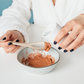 Hands pouring water into a bowl of pink clay facial mask powder while mixing with a ceramic spoon for a skincare ritual.