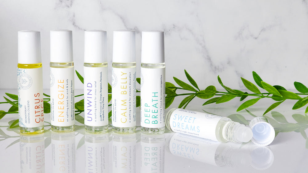A selection of six aromatherapy oil bottles labeled with different scents such as  Citrus Spice, Energize, Unwind, Calm Belly Deep Breath, and Sweet Dreams, displayed on a marble surface with green leaves in the background.