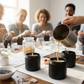 Bright, close-up view of hands pouring candle wax into glass jars during a candle-making workshop. Multi-racial, mixed-gender participants in the background, softly out of focus. Warm, clean lighting with natural daylight. Wooden tables, low profile black jars, wicks, dried botanicals, and linen accents. High-resolution lifestyle photography, cozy yet crisp.