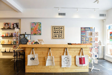 A retail shop interior with a light wood reception desk, white walls, and three canvas tote bags hanging from hooks on the front of the desk. A framed sign on the wall reads “SHOP MADE IN DC.