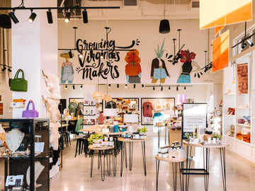 A bright, stylish boutique interior with display tables, shelves, and a large hand-painted wall mural reading 'Growing Virginias makers' with illustrated figures. The space has warm lighting, white walls, and multiple retail displays.