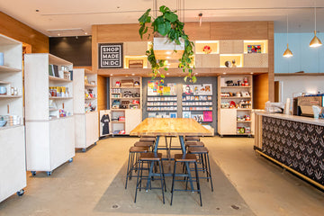 A bright, modern retail interior with a central wooden table and black bar stools, surrounded by white shelving units, a patterned counter, and a hanging green plant.