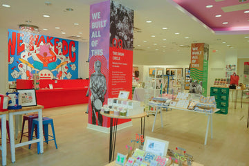 A bright, modern retail store interior with colorful display tables, posters, and wall art. A large central pink and red pillar reads 'WE BUILT ALL OF THIS' with a black-and-white photo, and a blue mural on the left says 'WE WAKE DC'.