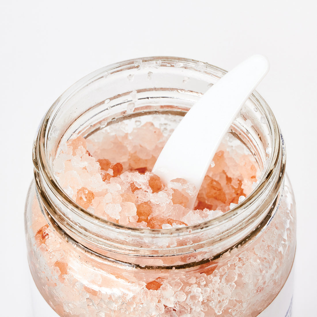 A glass jar of Calm Unwind + Relax Bath Soak filled with pink salt crystals, set on a stone surface next to a folded white towel and a small wooden scoop, with a softly blurred bathroom background.