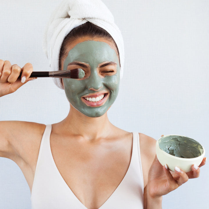Smiling woman having a facial treatment with the detoxifying green clay face mask on her face. Hello Wellness.